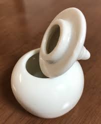 A round, white ceramic pot with a pointed lid rested on its side so that the opening of the pot is visible sits on a wooden surface. There are concentric rings designed into the lid.